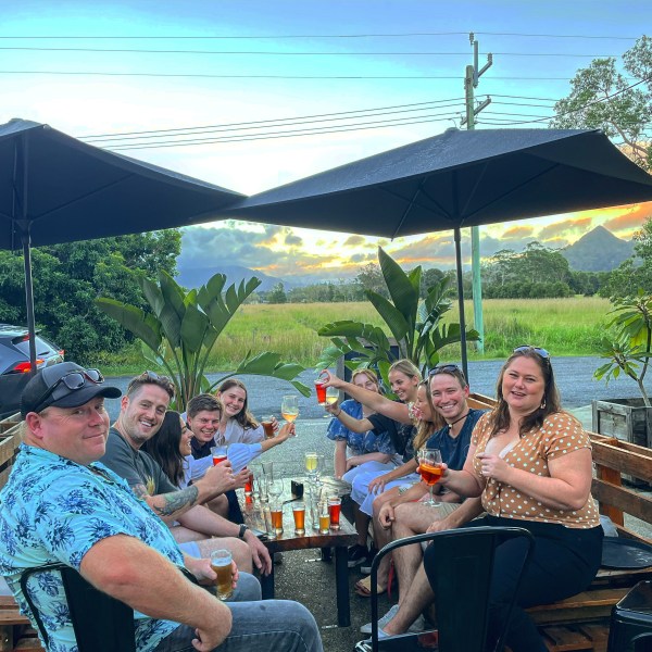 Group of people toasting with drinks at an outdoor bar under umbrellas during sunset.