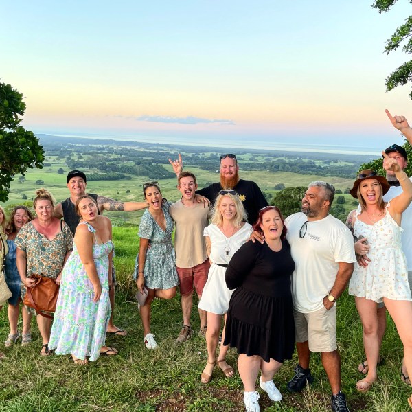 Group of people smiling and posing on a grassy hill with a scenic view of the countryside and sky.
