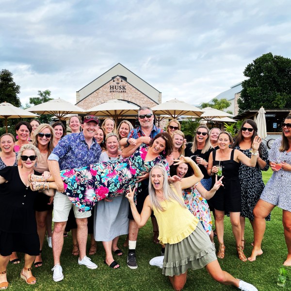 Group of people outdoors lifting one person, smiling and posing playfully, with a building in the background.