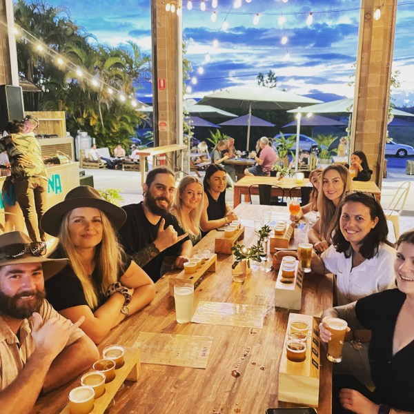 Group of people seated at a wooden table with drinks, under string lights in an outdoor setting.