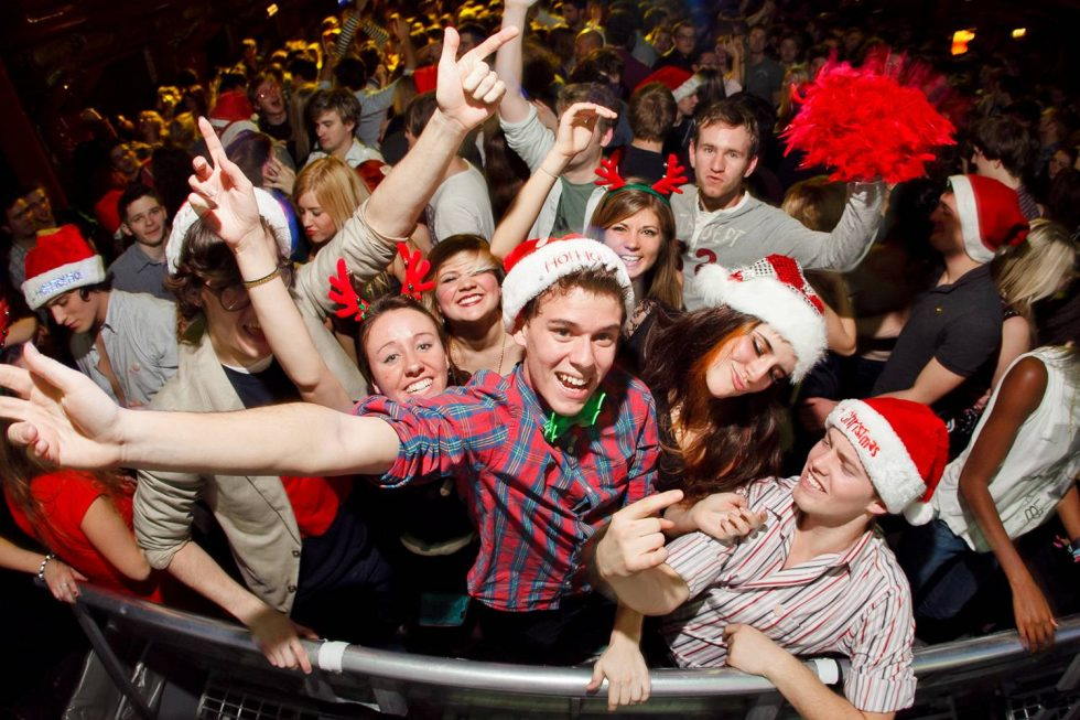 Crowd of people at a party wearing Santa hats and reindeer antlers, smiling and cheering.