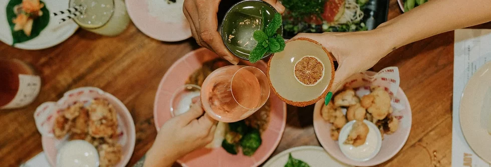 Hands toasting with cocktails over a table of food dishes.