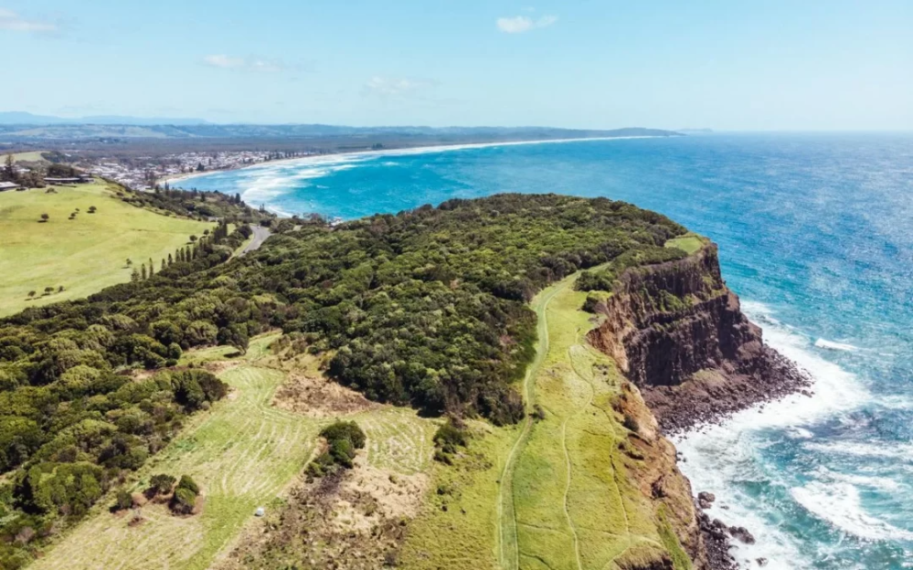 Aerial view of a coastal cliff with lush greenery, ocean waves and a distant town.