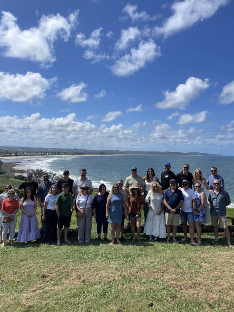 Group of people standing on grass with ocean and partly cloudy sky in background.